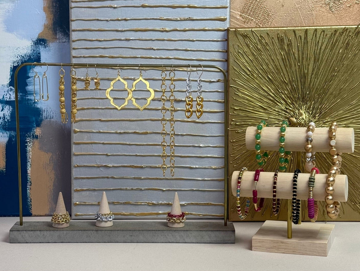 A styled jewelry display setup featuring a gold metal earring stand on a gray base, a vertical striped board with gold horizontal bars, and a wooden double bracelet holder with colorful beaded bracelets, all arranged against an abstract blue, white, and gold background.