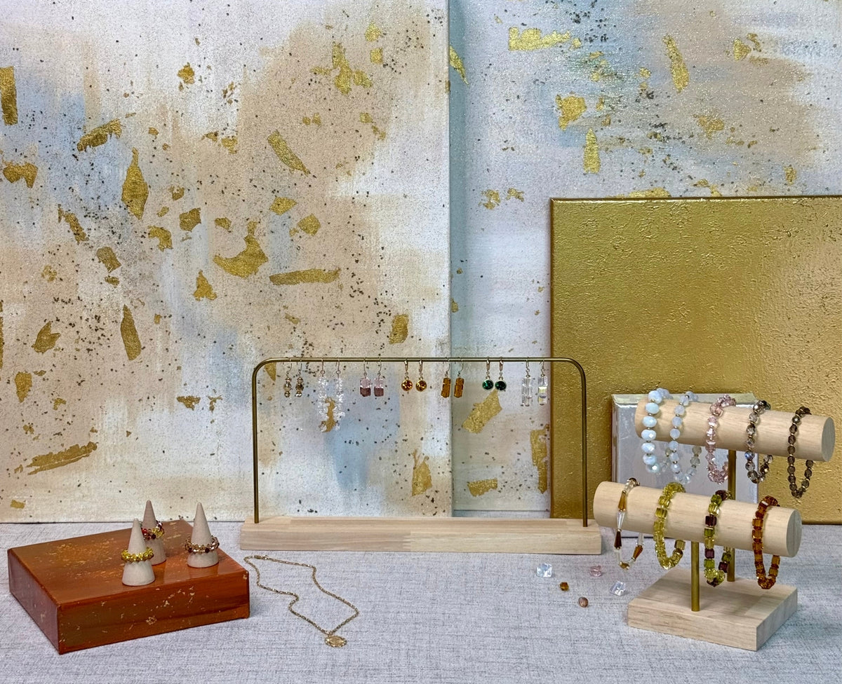 A styled jewelry display featuring multiple necklaces and bracelets on wooden rolls, a gold-framed necklace stand, and a small wooden box with decorative pieces, set against an abstract gold, blue, and white textured wall.