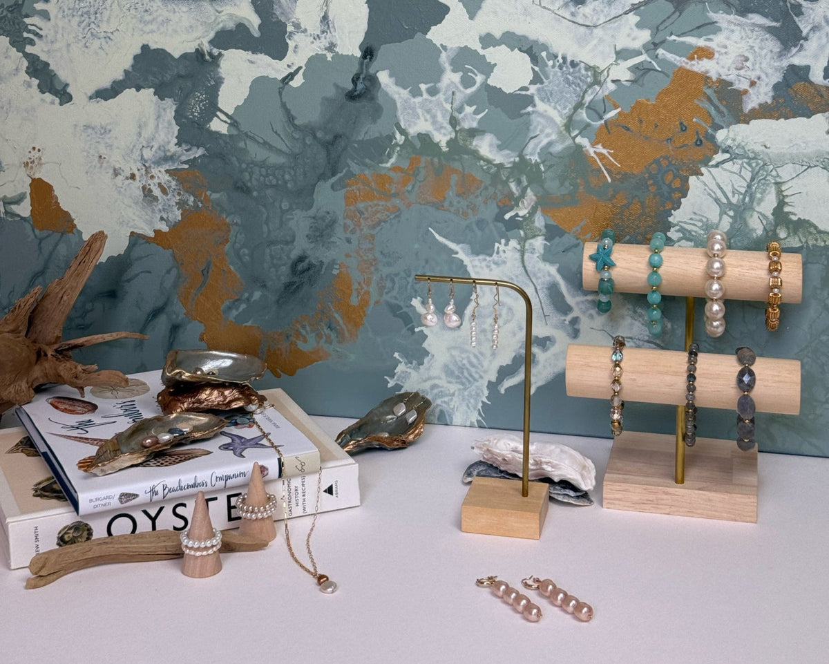 A styled jewelry display on a white tabletop against a large abstract wall mural in muted blue, gray, white, and gold tones. The display includes wooden earrings stands holding bracelets and necklaces, a gold earring stand with dangling earrings, and scattered jewelry pieces.