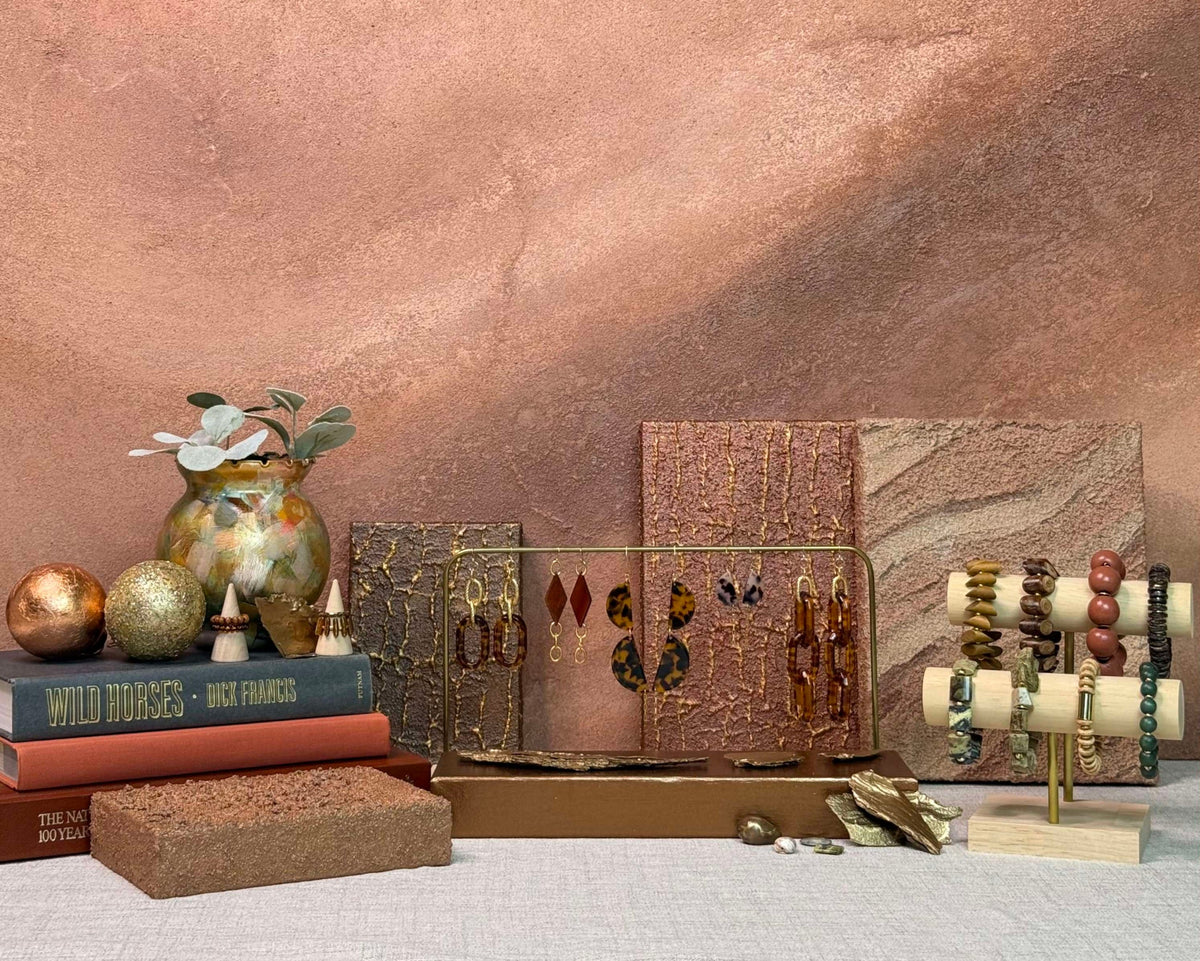 A styled jewelry display featuring multiple earrings and bracelets on stands, arranged with decorative books, a vase, and textured stone-like panels against a warm copper-toned backdrop.